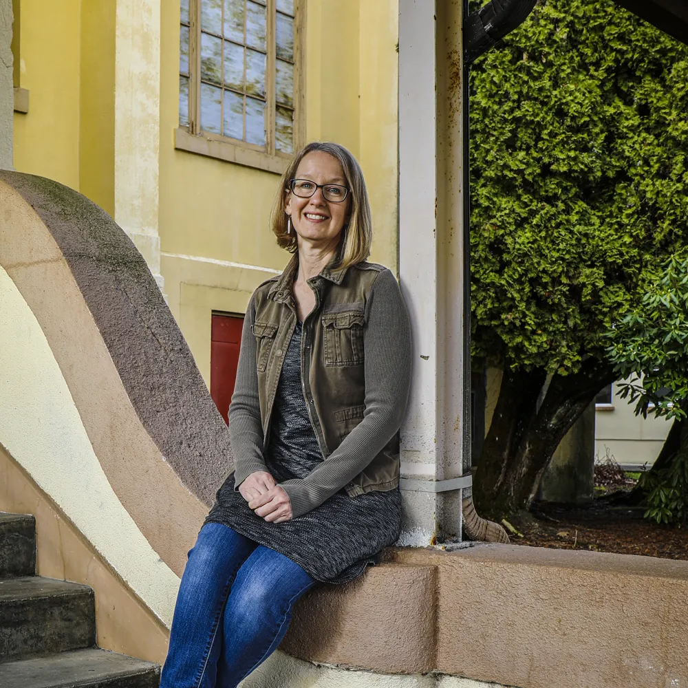 Smiling woman with glasses sitting on outdoor ledge near stairs and trees.