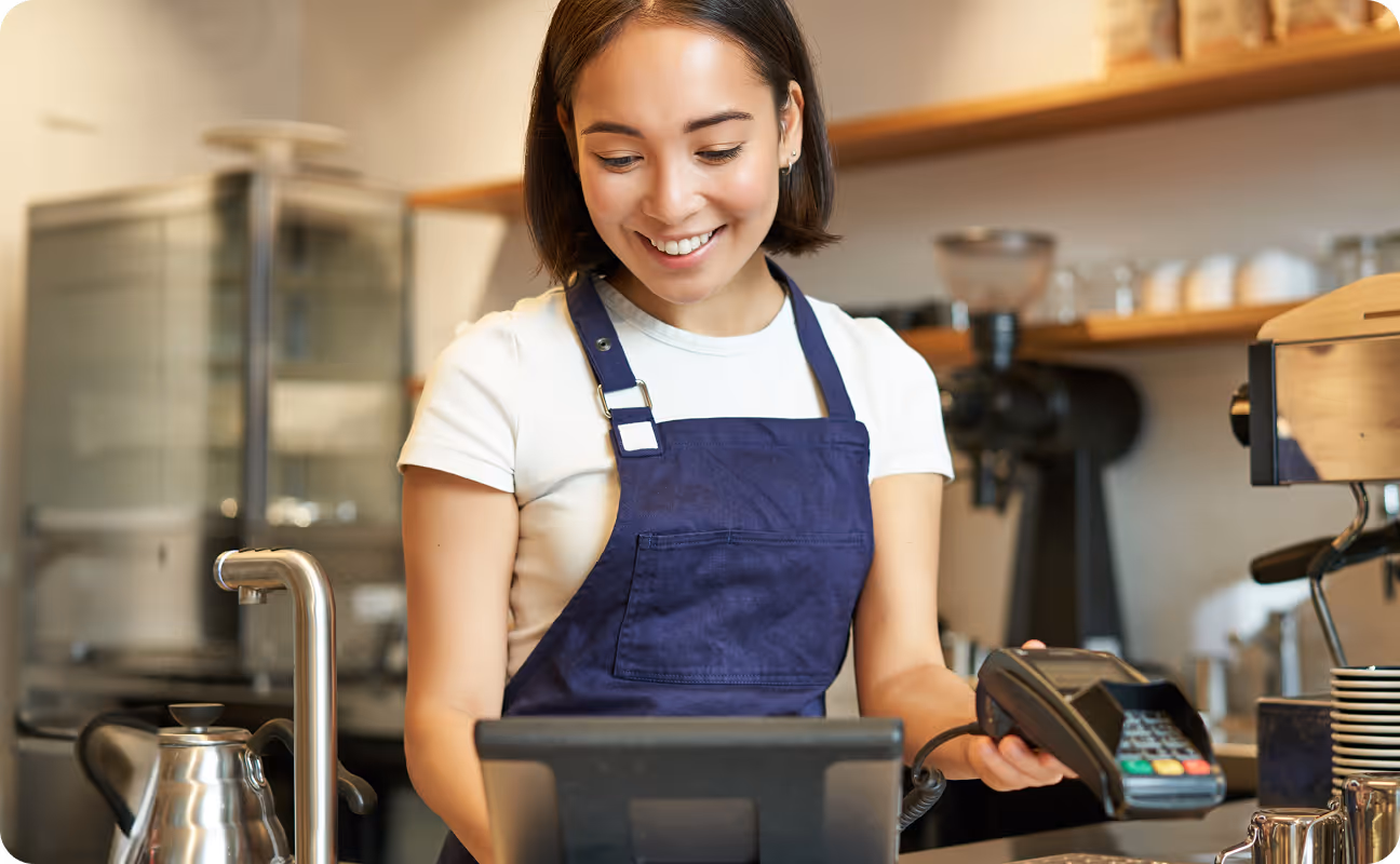 Smiling barista in a purple apron holding a card reader at a coffee shop register.