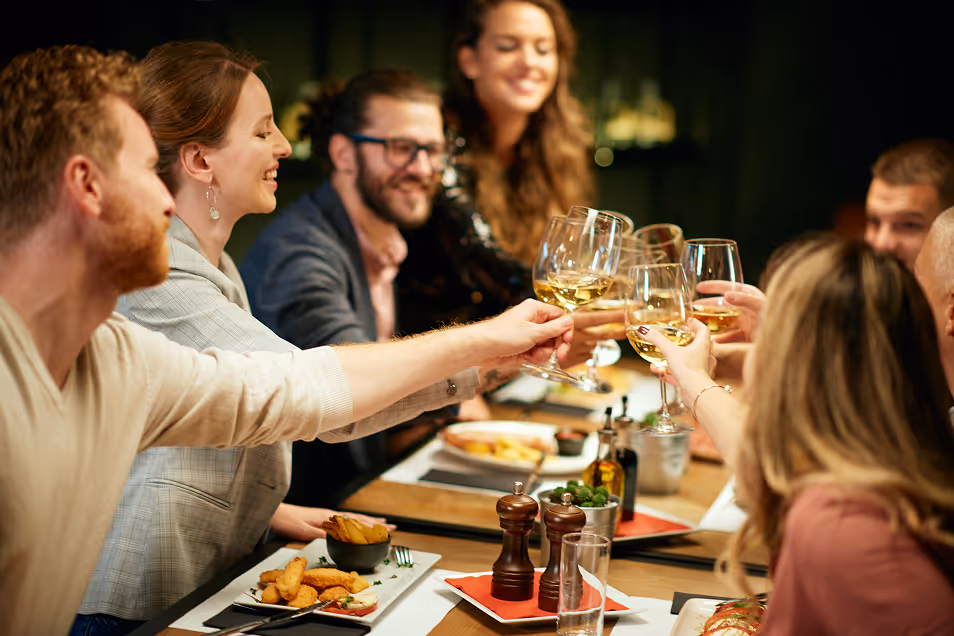 Group of friends clinking glasses with white wine at a dinner table with food and condiments.