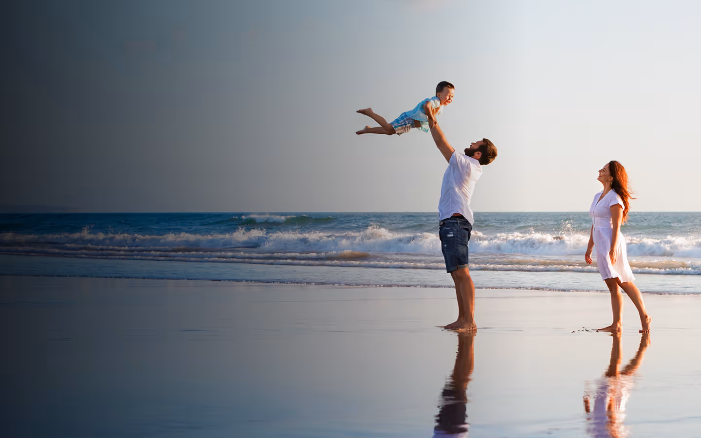 Man lifting a happy child in the air on a beach with waves and woman walking nearby.