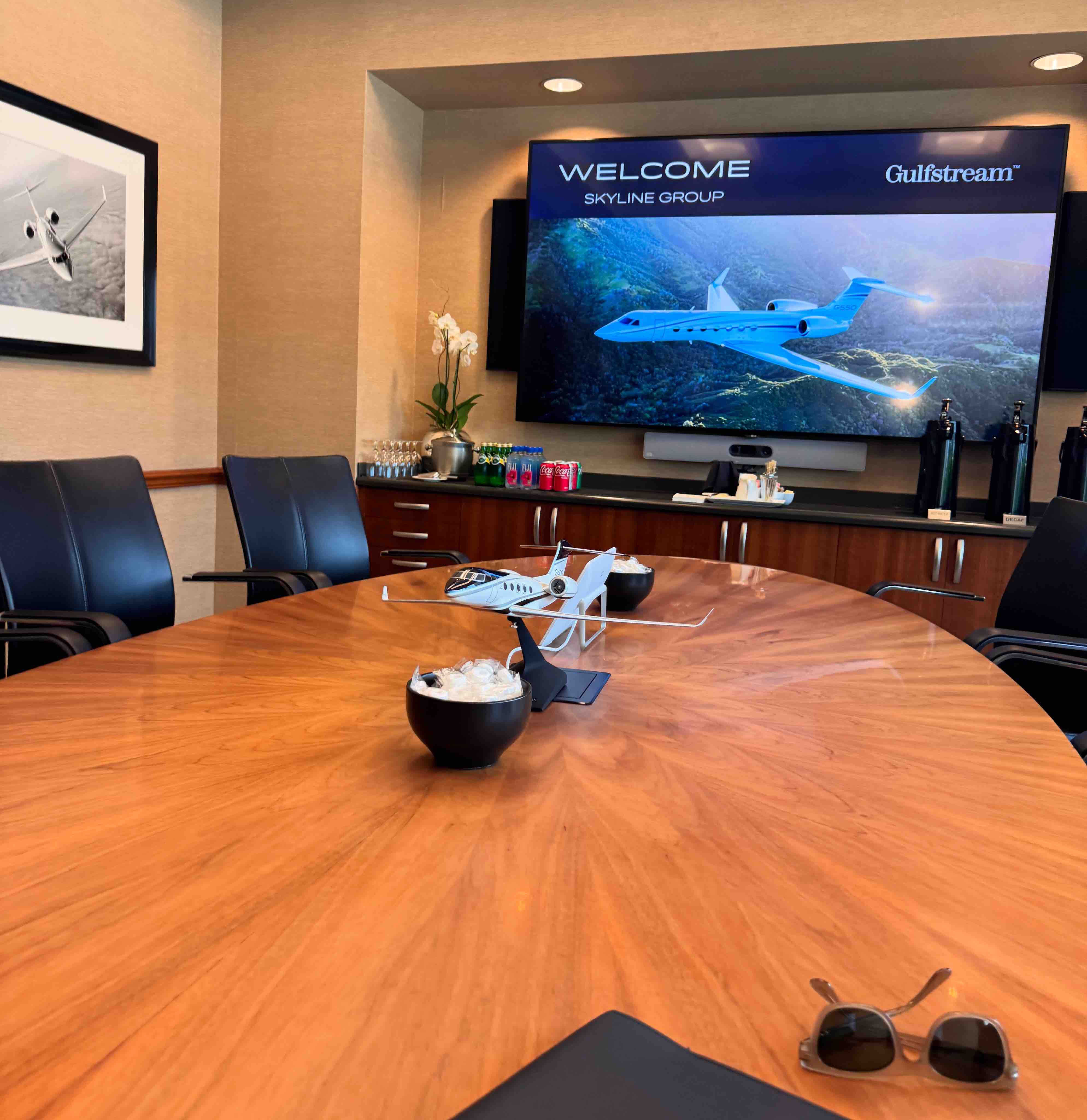 Conference room with wooden table, model airplane, sunglasses, chairs, and screen displaying Gulfstream welcome.
