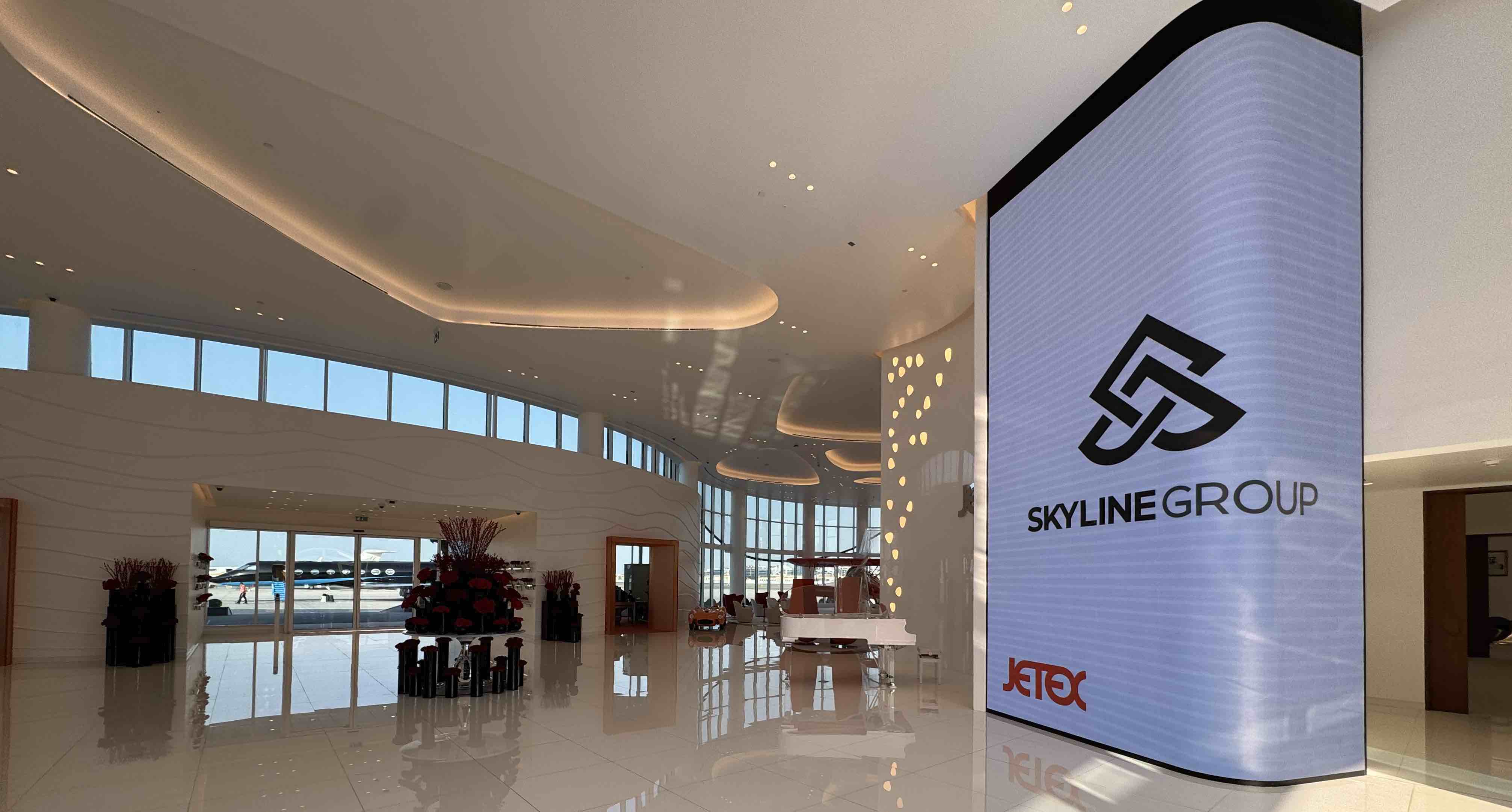 Modern airport lounge with large digital Skyline Group sign and airplane visible outside glass doors.