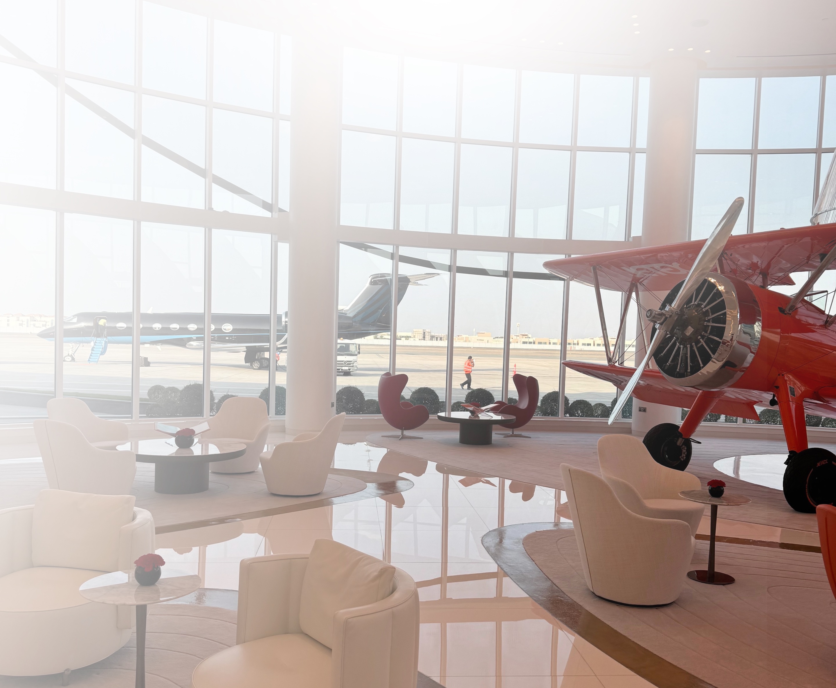 Modern airport lounge with white chairs, small tables, a red vintage airplane, and a private jet outside large windows.