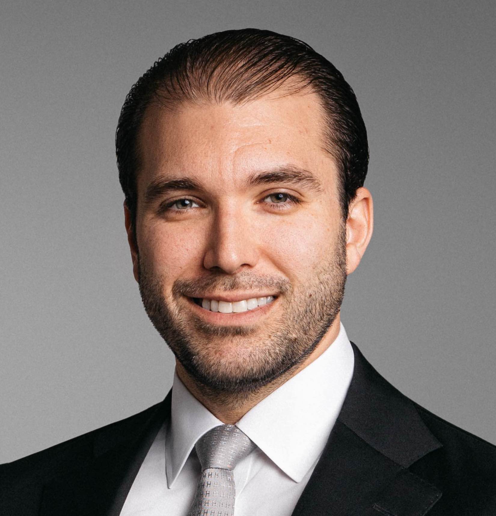 Smiling man with short dark hair and beard wearing a black suit, white shirt, and silver tie.
