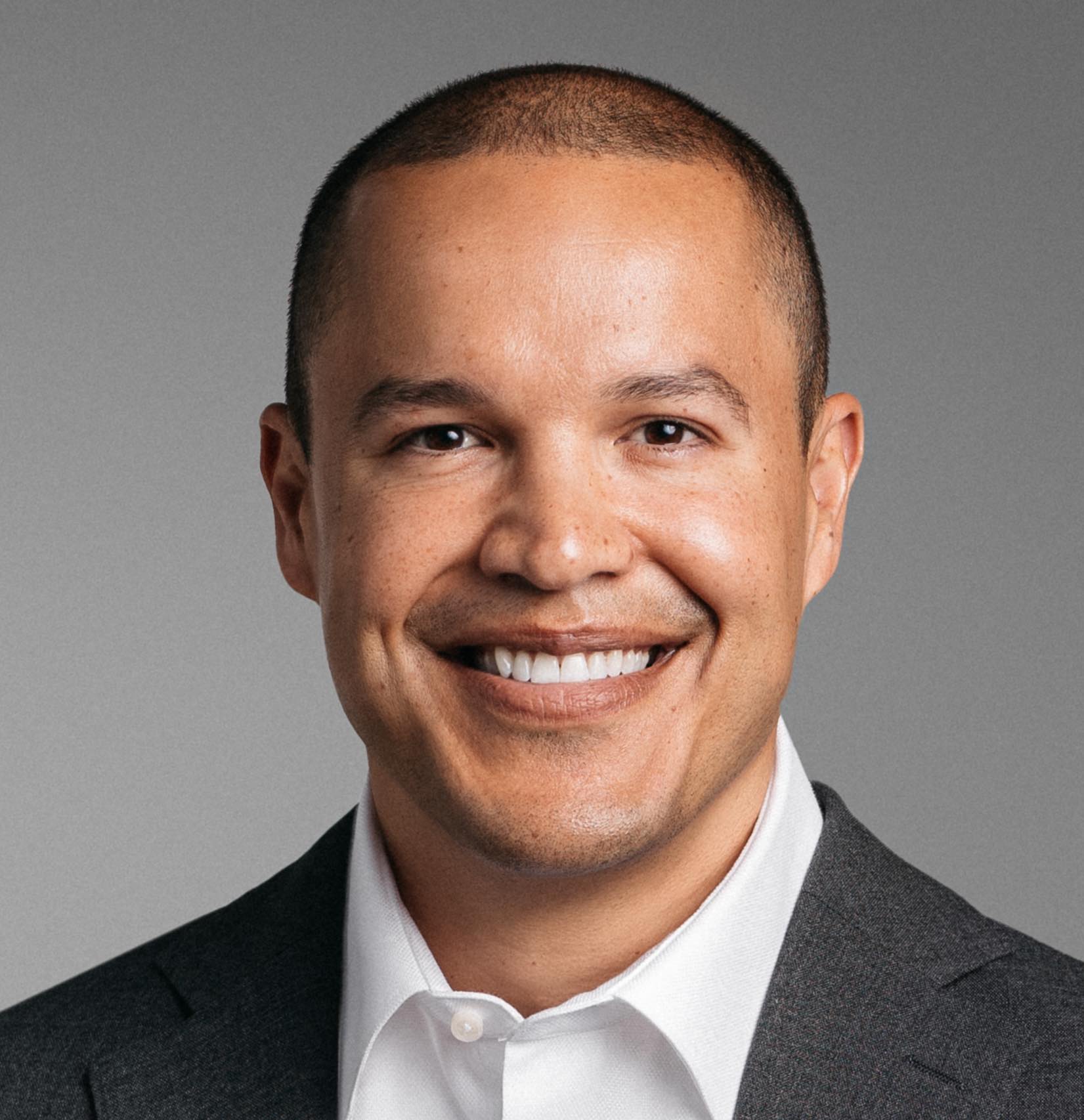 Smiling man with short hair wearing a white shirt and dark blazer against a gray background.