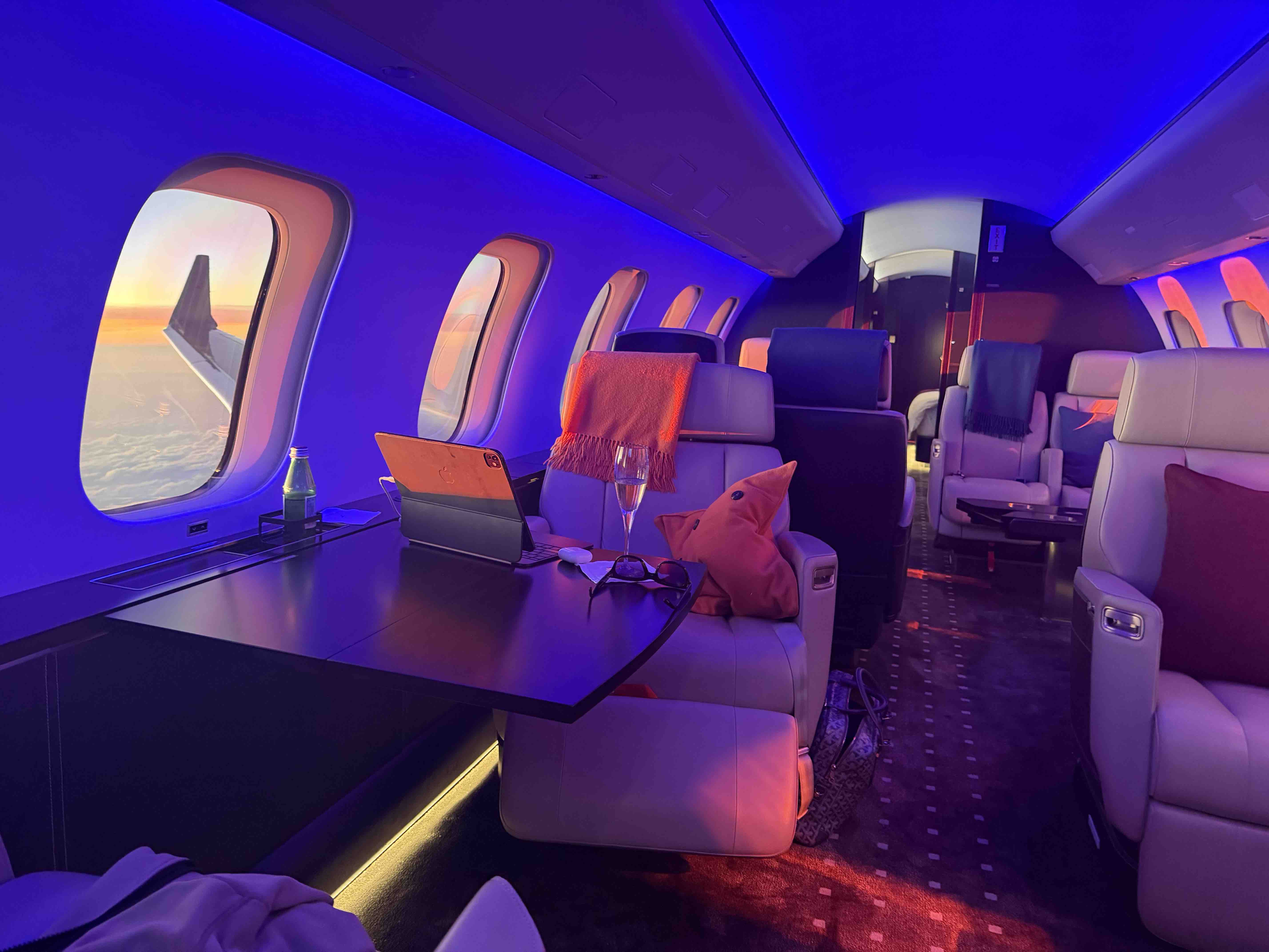 Interior of a private jet with leather seats, a tablet, drink, and sunset glowing through the windows.
