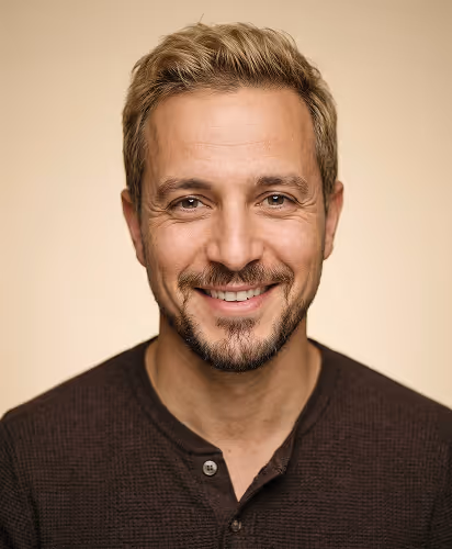 Smiling man with short blond hair, beard, light brown eyes, wearing a dark brown shirt on beige background.