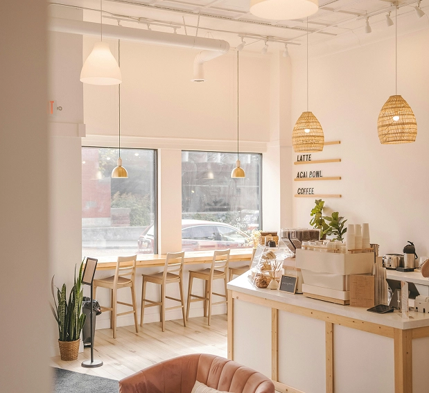 Bright cozy coffee shop with wooden chairs, pendant lights, a service counter, and potted plants.