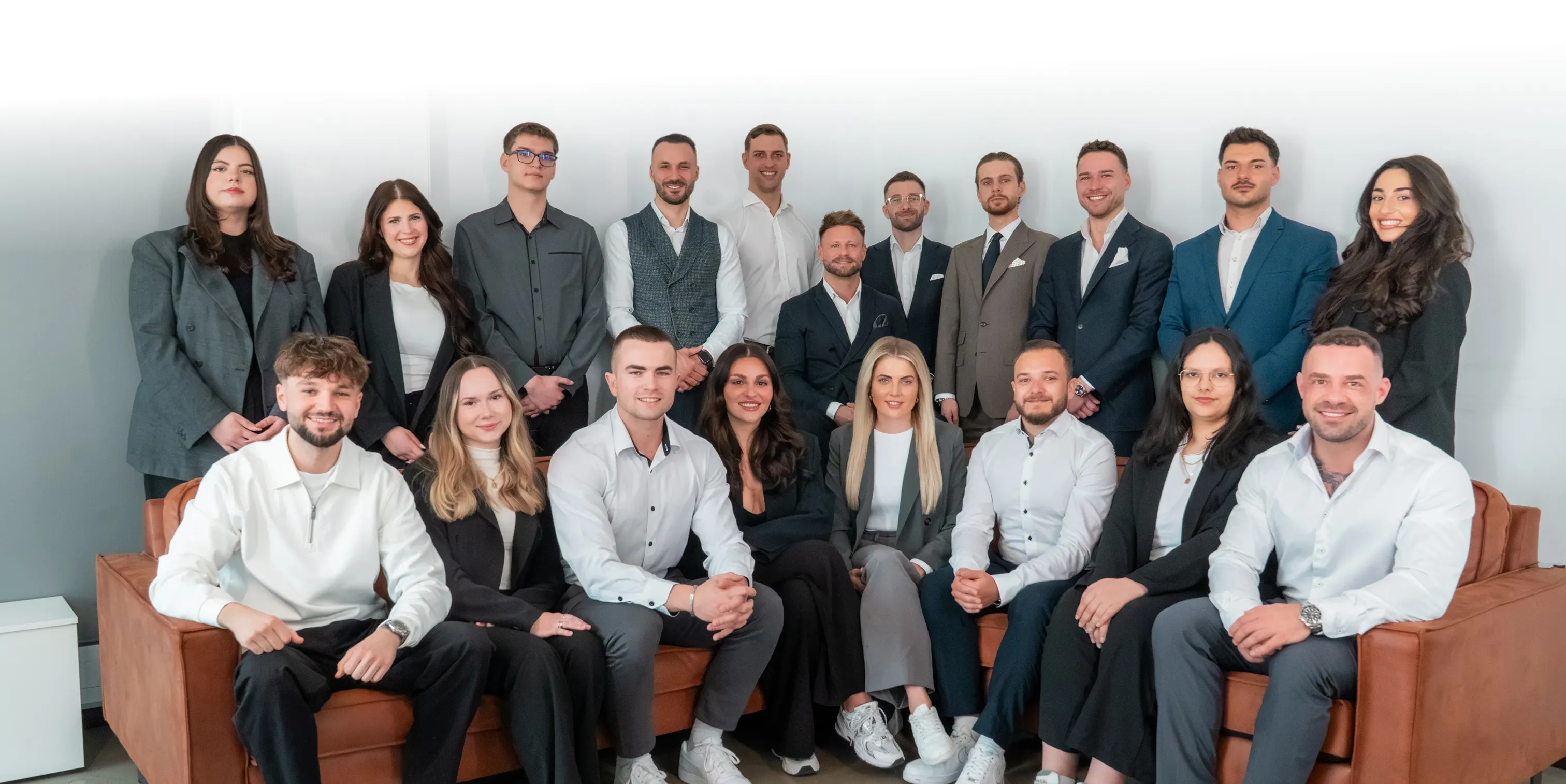 Gruppenfoto von 19 jungen, professionell gekleideten Menschen in einem hellen Büro vor weißem Hintergrund.