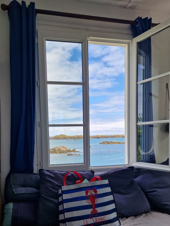 Vue sur une fenêtre ouverte sur la mer avec des coussins bleus et un sac rayé à motif homard.