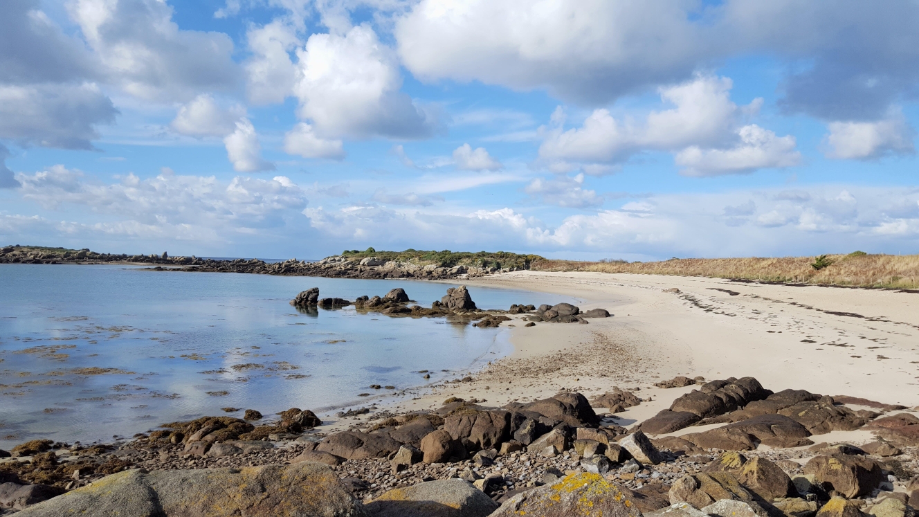 Une plage de sable clair avec des rochers et une mer calme sous un ciel partiellement nuageux.