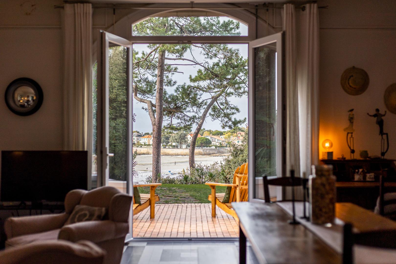 Vue de l'intérieur d'un salon avec portes ouvertes donnant sur une terrasse, jardin et rivière.