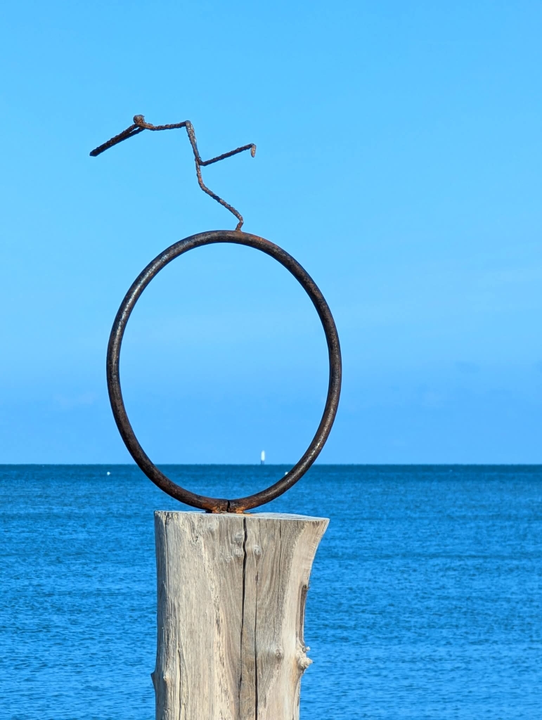 Sculpture métal ronde sur socle en bois face à la mer avec ciel bleu clair en arrière-plan.