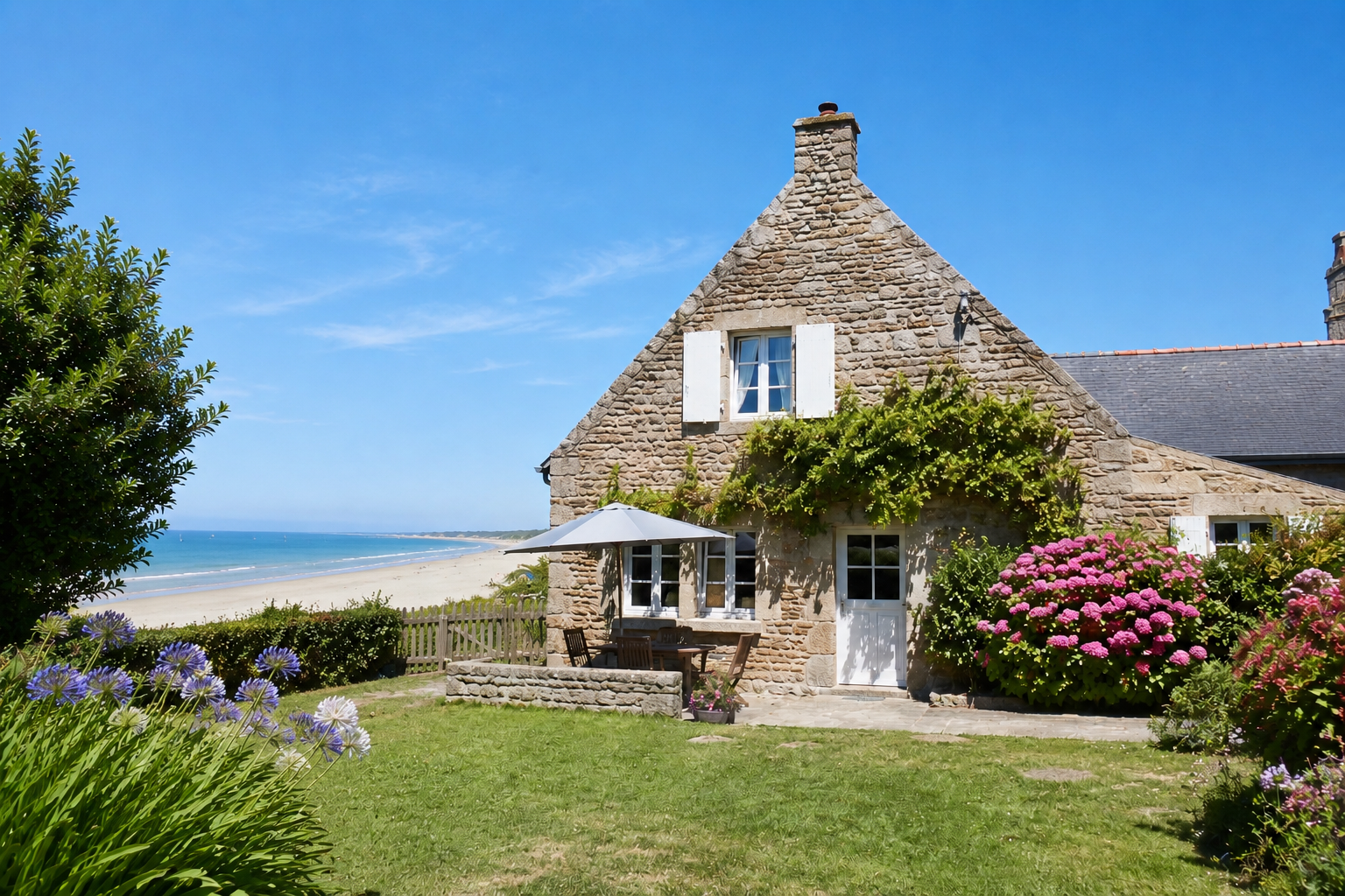 Maison en pierre avec jardin fleuri, parasol, vue sur plage et océan sous ciel bleu.