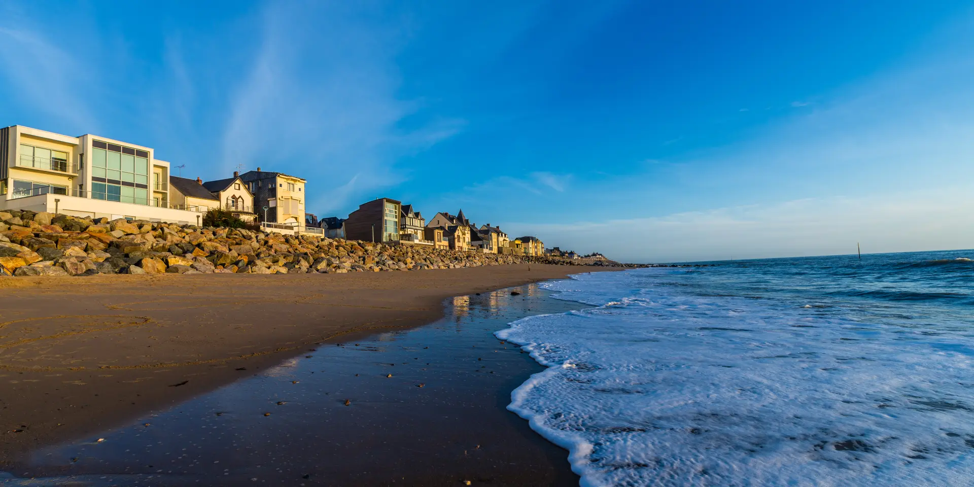 Maisons alignées sur une plage rocheuse avec sable et océan sous un ciel bleu clair.