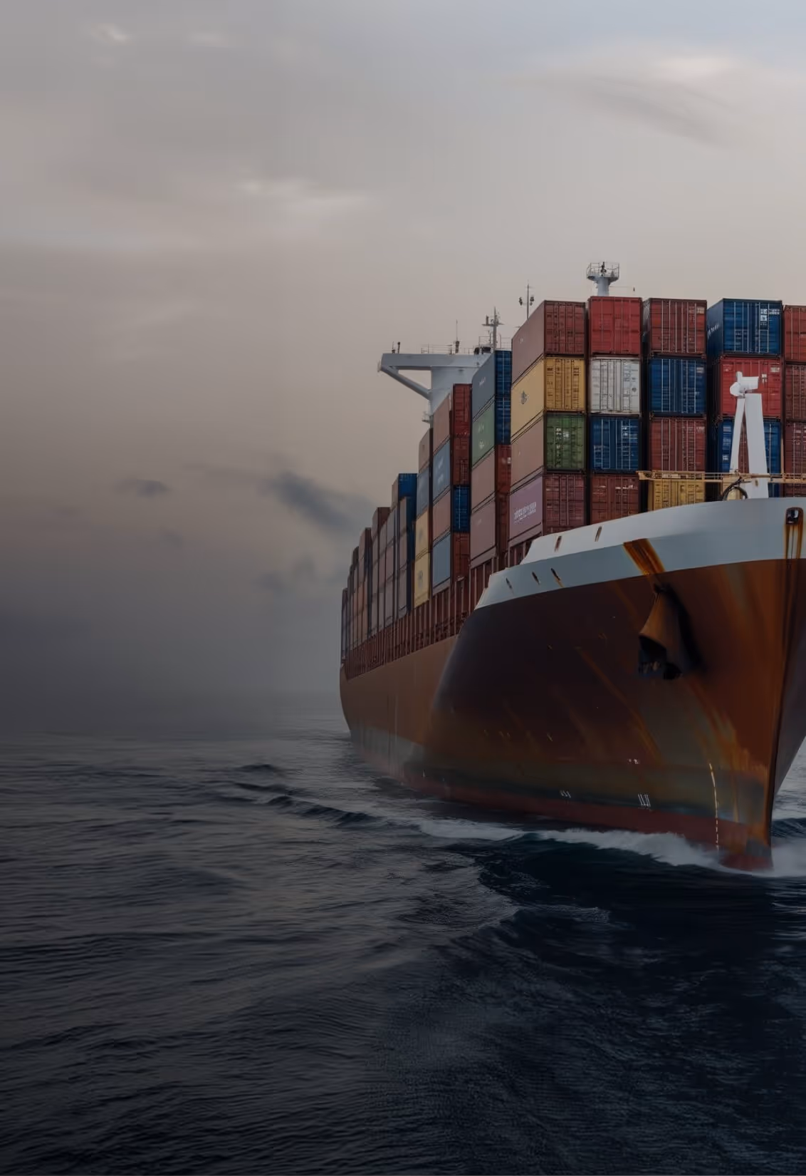 Cargo ship loaded with multicolored shipping containers sailing on dark ocean under a cloudy sky.