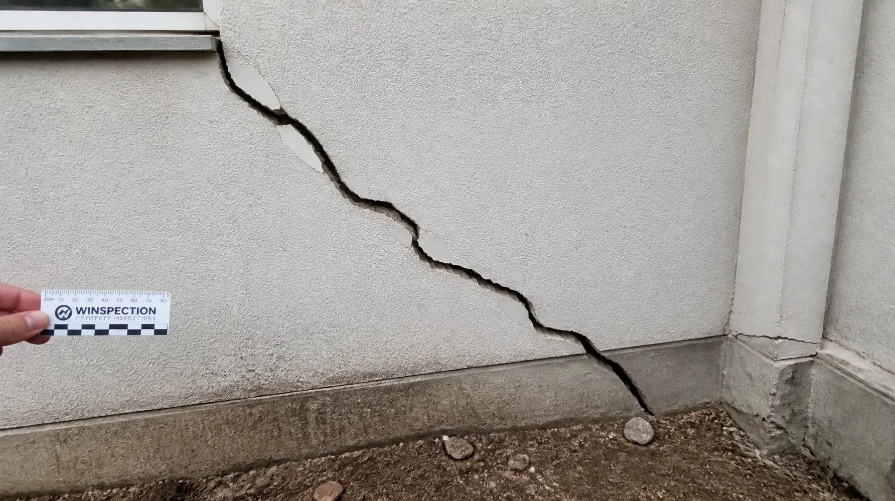 A significant diagonal crack on an external rendered wall, assessed with a measuring scale for forensic reporting.
