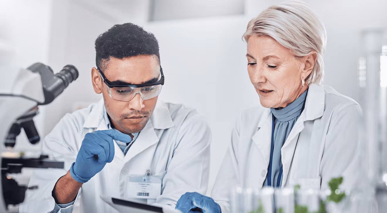 Two scientists in lab coats and gloves examining a sample or data on a tablet in a lab.