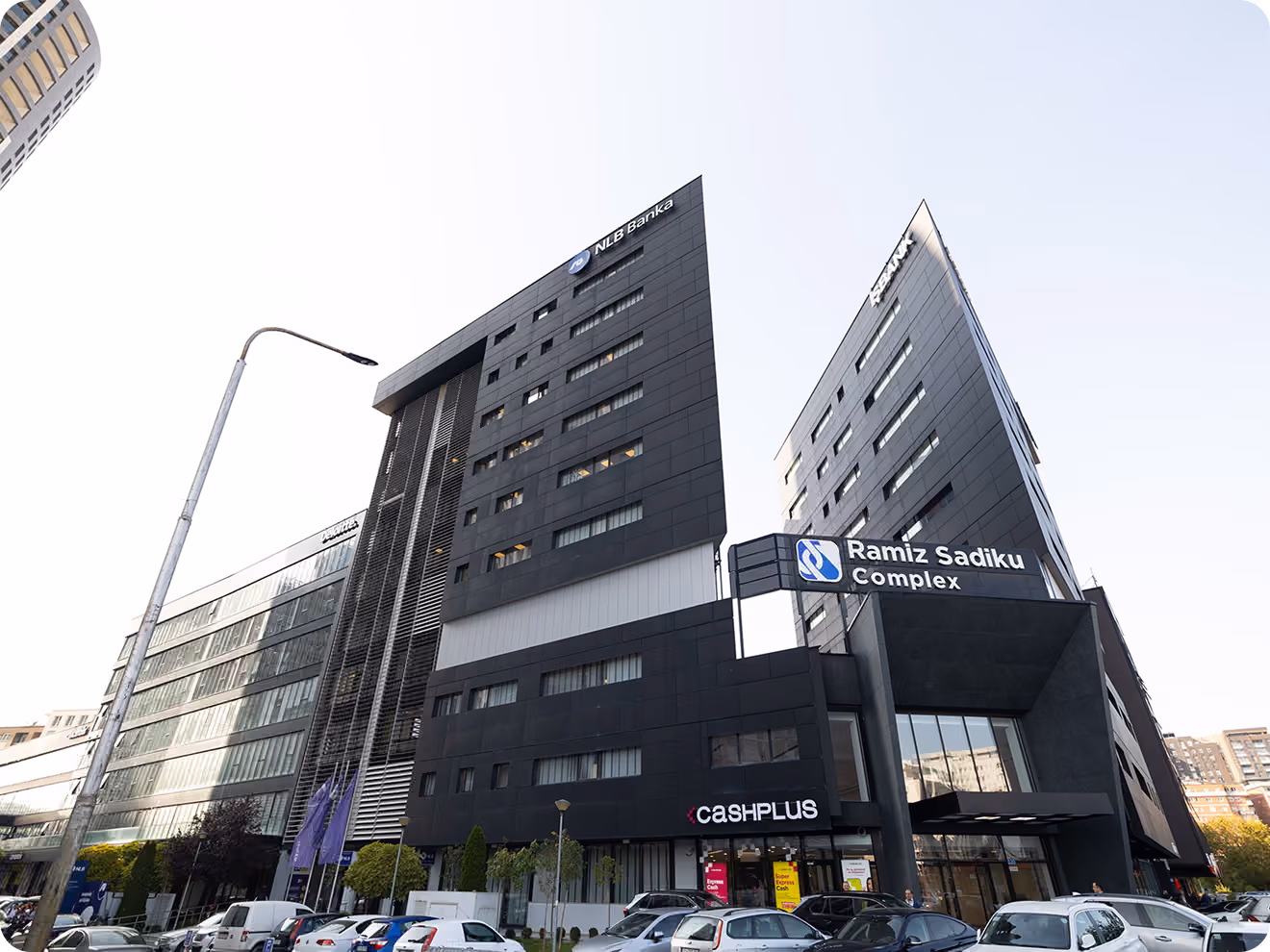 Modern multi-story office buildings including NLB Bank and Ramiz Sadiku Complex with cars parked in front under a clear sky.