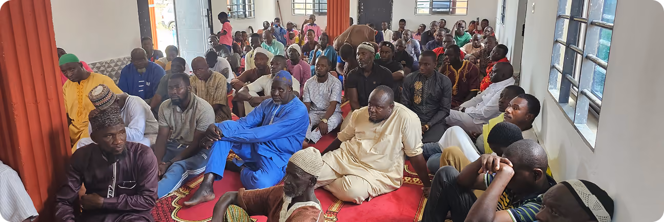 Men gathered on prayer mats inside a mosque hall in Africa, listening attentively during a community gathering.