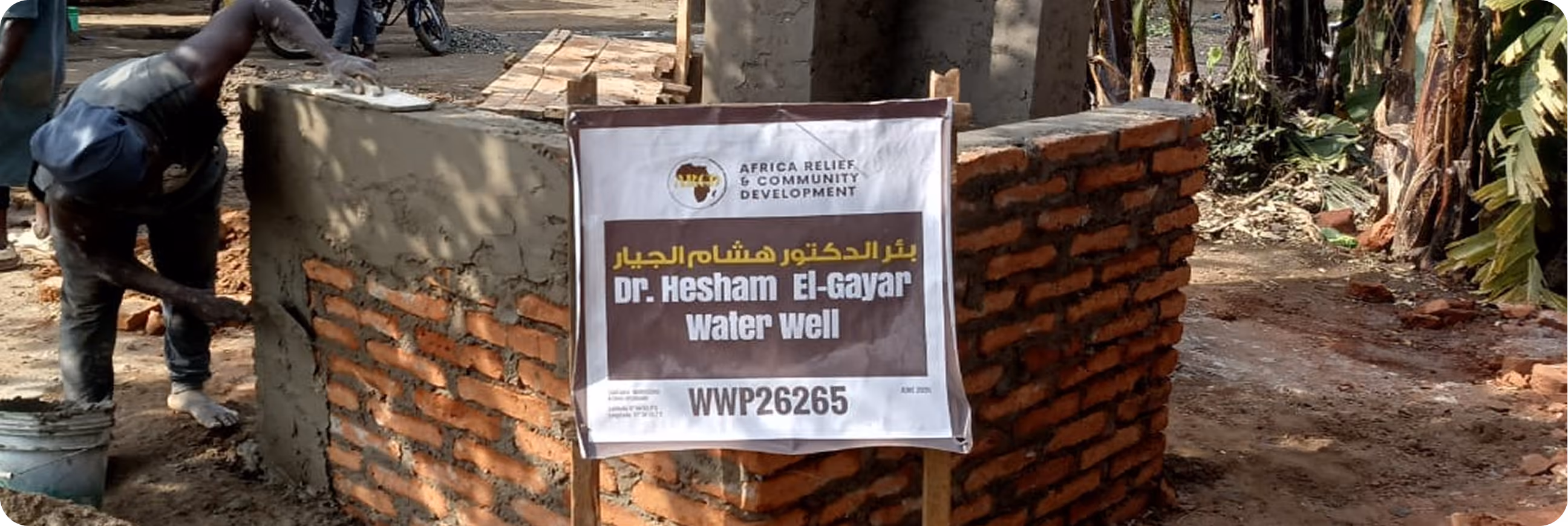 A worker plasters a brick well casing beside an ARCD dedication sign for the Dr. Hesham El-Gayar Water Well.