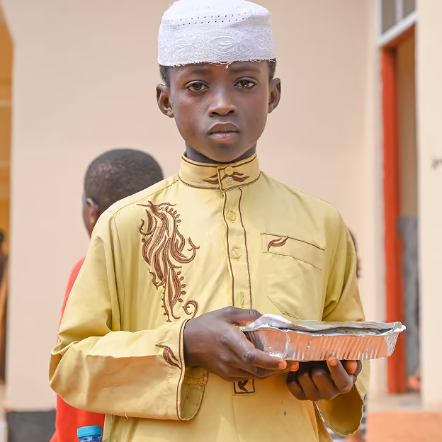 A young boy wearing a light yellow embroidered tunic and white embroidered cap holding a foil meal container with both hands.