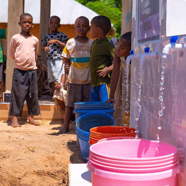 Child drinking water from a metal hand pump over muddy ground with a yellow container nearby