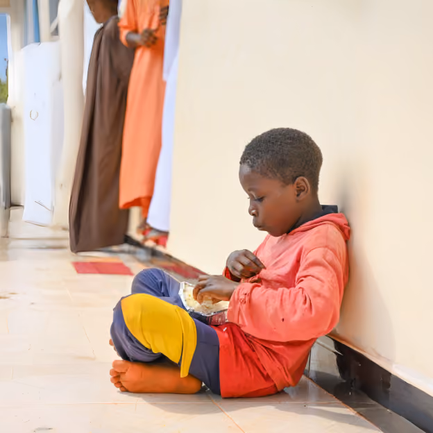 Young child sitting on the floor beside a wall and eating a meal from a small container.