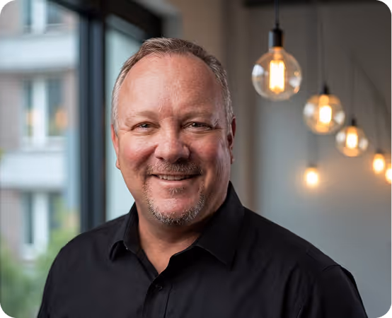 Smiling middle-aged man in black shirt standing indoors with hanging light bulbs in background.