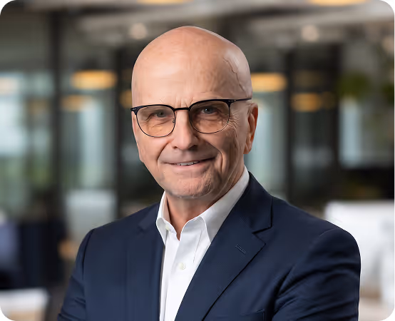 Bald man in glasses wearing a dark blazer and white shirt, smiling in an office setting.