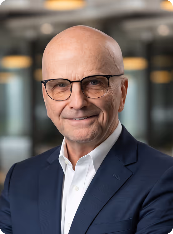 Portrait of a bald man with glasses in a dark suit and white shirt smiling indoors.
