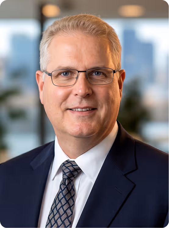 Middle-aged man with glasses wearing a dark suit, white shirt, and patterned tie, smiling indoors.