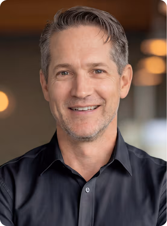Smiling man with short gray hair wearing a dark button-up shirt in a softly lit indoor setting.