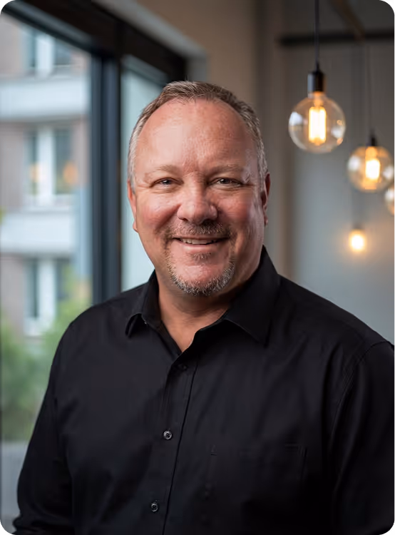 Smiling middle-aged man with short hair and goatee wearing a black shirt indoors near windows and hanging lights.