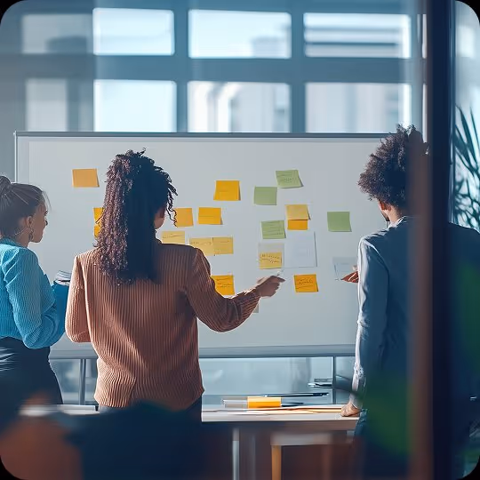 Three people stand facing a whiteboard covered in orange and green sticky notes in a bright office.