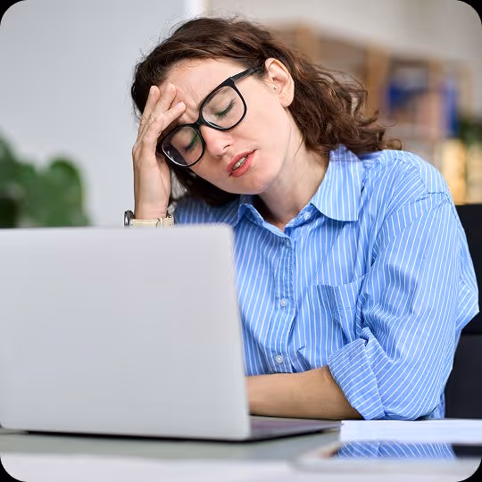 Tired woman with glasses in blue shirt resting head on hand while sitting at a laptop.