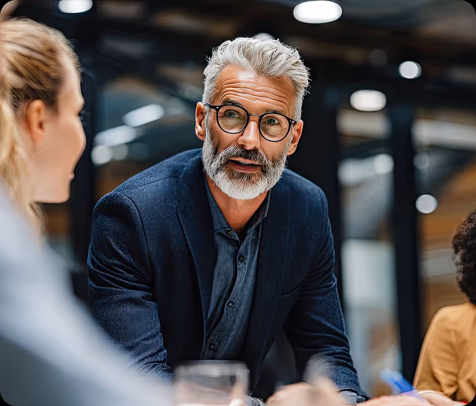 Older man with gray hair and beard wearing glasses and a blazer talks to a colleague in an office.