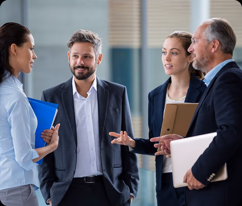 Four professionals in business attire having a discussion in an office setting, holding folders and laptop.