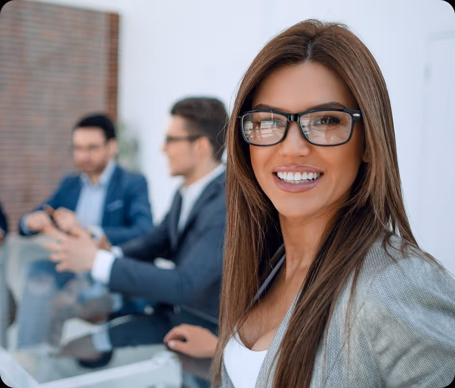Smiling businesswoman wearing glasses and a blazer with two blurred men in suits in the background.