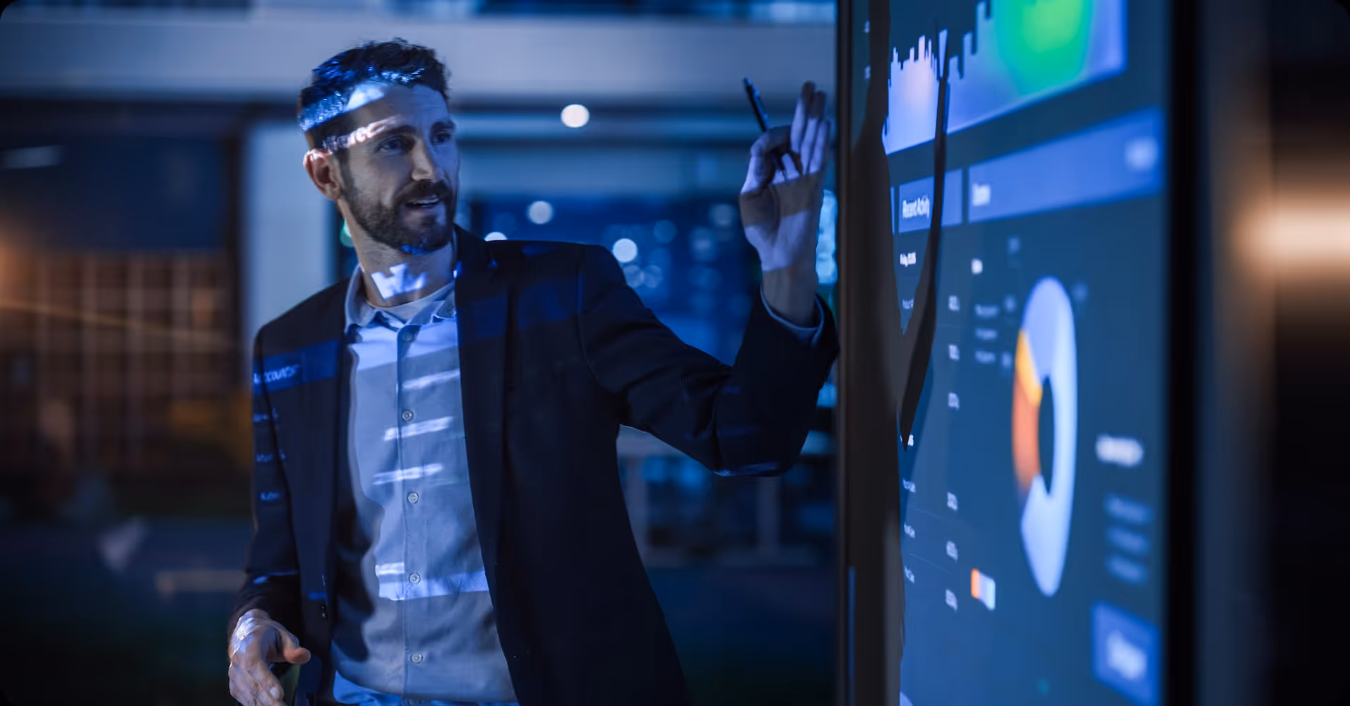 Man in suit using digital touch screen with charts and graphs in dark modern office.