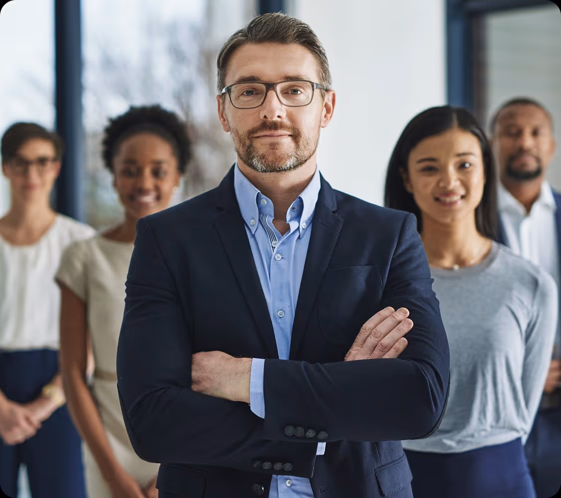 Confident man in glasses and navy suit stands with crossed arms, team of diverse professionals behind him.