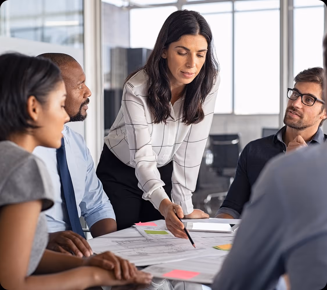 Businesswoman points at documents on table while colleagues listen attentively in office meeting.