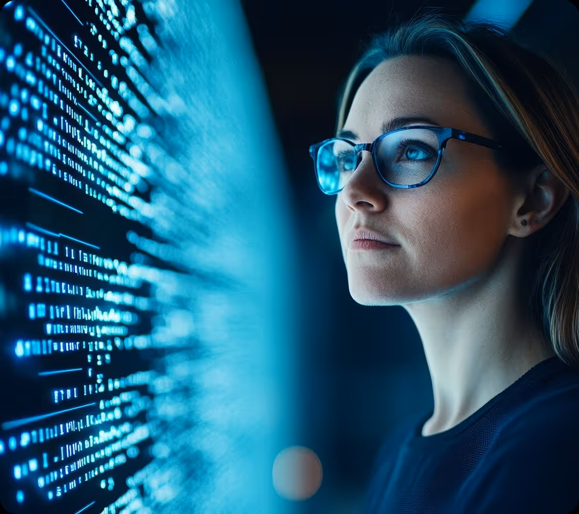 Young woman with glasses studies blue glowing code on a screen in a dark environment.