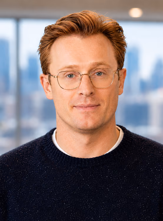 Portrait of a man with red hair, glasses, and a dark sweater, with a cityscape blurred in the background.