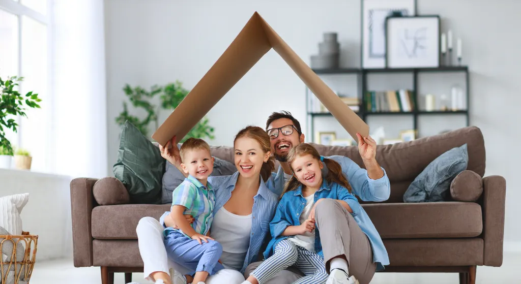 Smiling family of four sitting on floor with cardboard roof symbolizing home protection