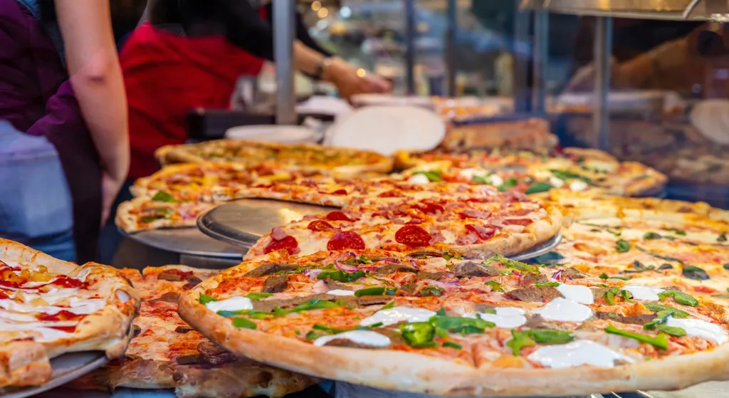 Multiple large pizzas with various toppings displayed in a pizzeria behind a glass case.