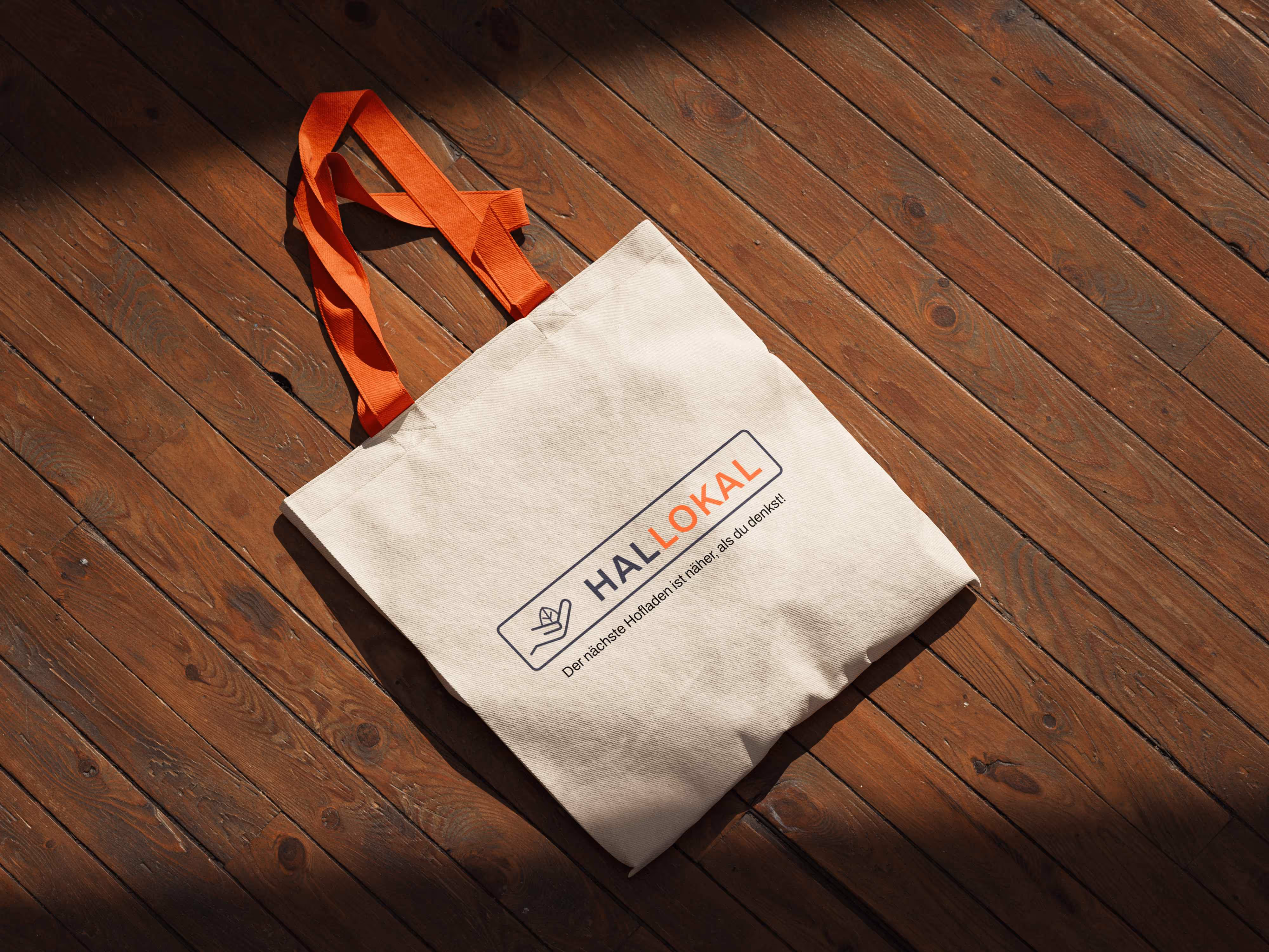 Stofftasche mit orangen Griffen und Aufdruck 'HalloLokal' auf Holzboden im Sonnenlicht.