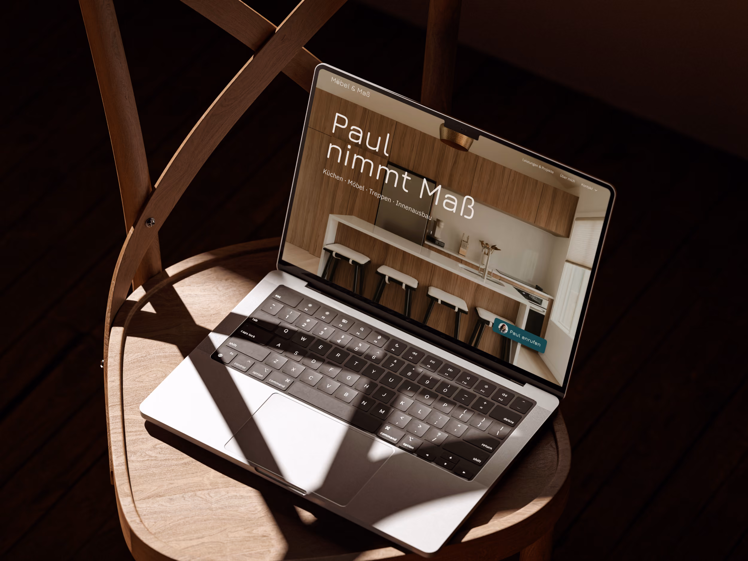 Laptop auf einem runden Holzstuhl mit einer Webseite, die eine moderne Küche mit Barhockern und Schränken zeigt, auf dem Bildschirm. Der Text auf der Webseite lautet: ‚Paul nimmt Maß‘, mit Untertiteln für Küchen, Möbel, Treppen und Innenausbau.