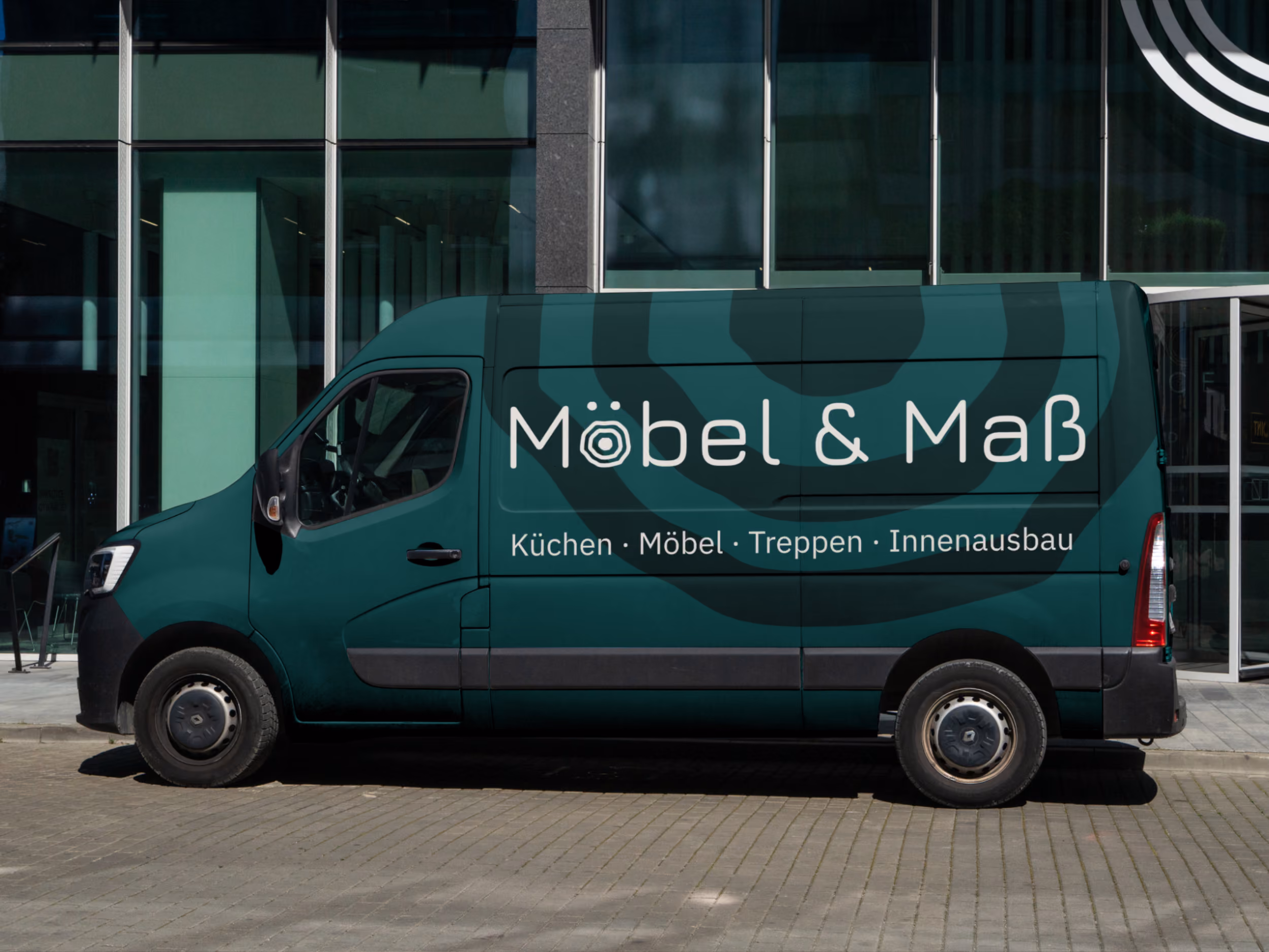 Dunkelgrüner Transporter mit der Aufschrift Möbel & Maß, darunter Küchen, Möbel, Treppen, Innenausbau, geparkt vor einem modernen Glasgebäude.