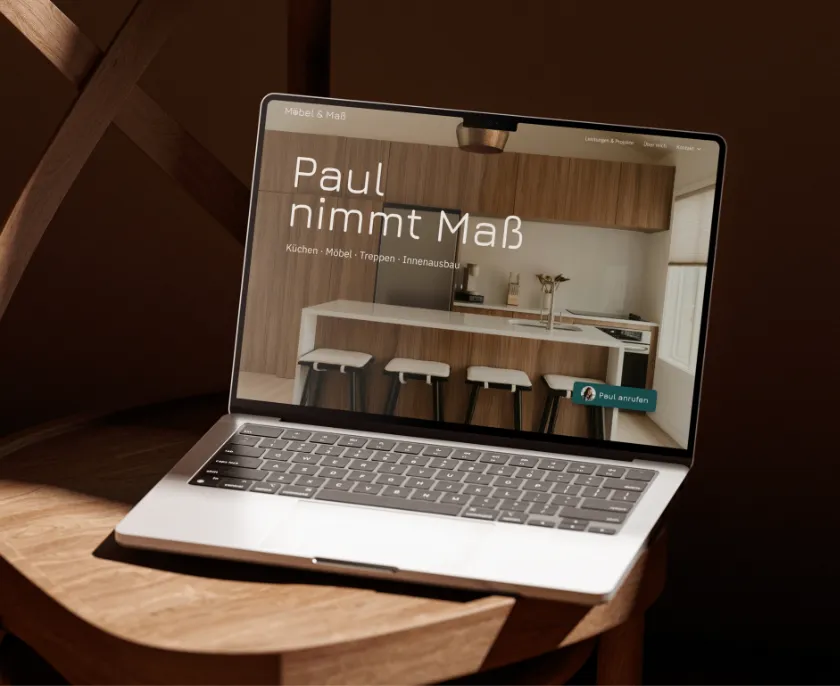 Ein Laptop auf einem Holzstuhl zeigt eine Webseite mit der Überschrift 'Paul nimmt Maß' und Bildern einer modernen Küche mit Holzschränken und weißen Barhockern.